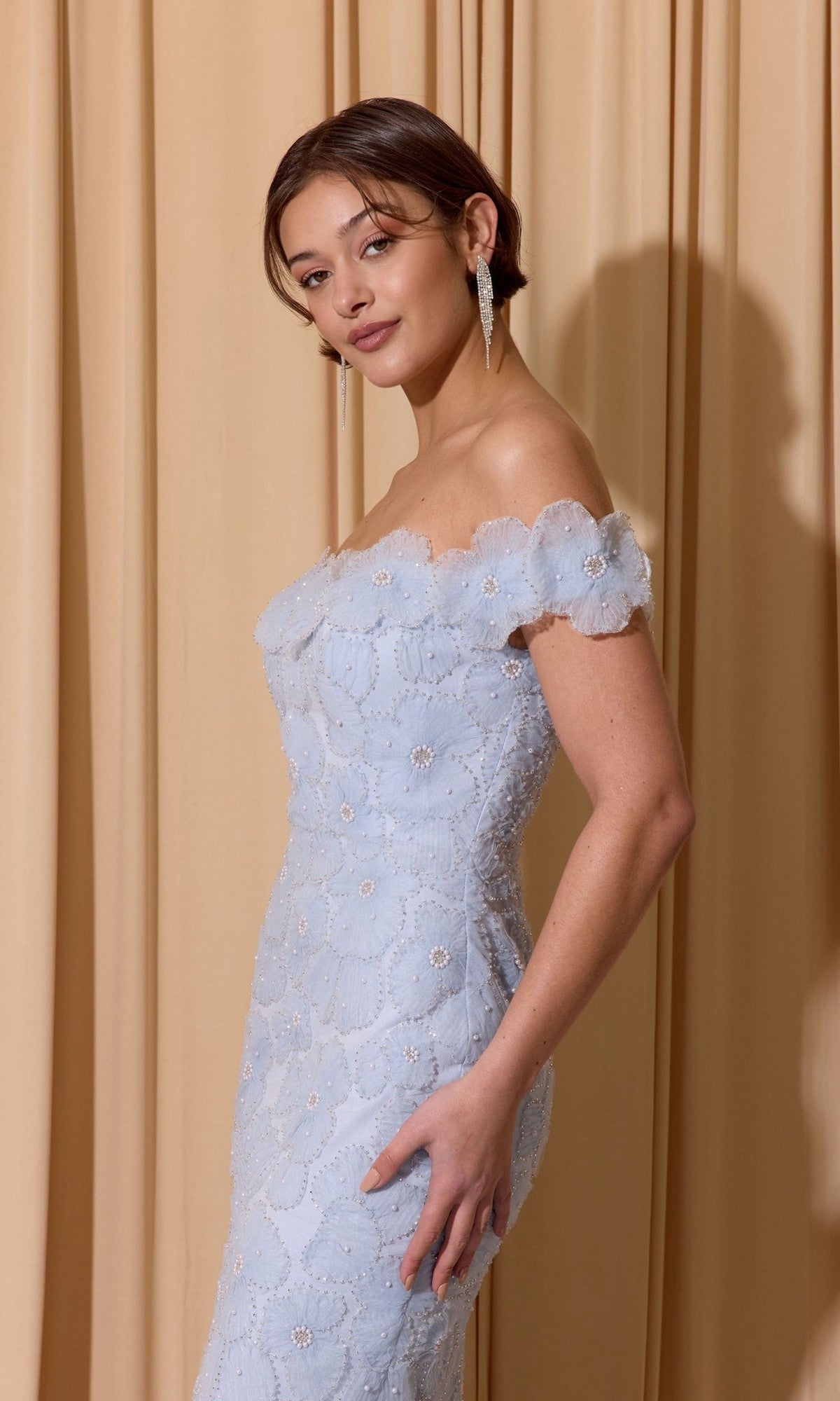 A woman in the Eureka EK205 long prom dress with light blue embroidery poses before beige curtains, wearing drop earrings and styled hair.